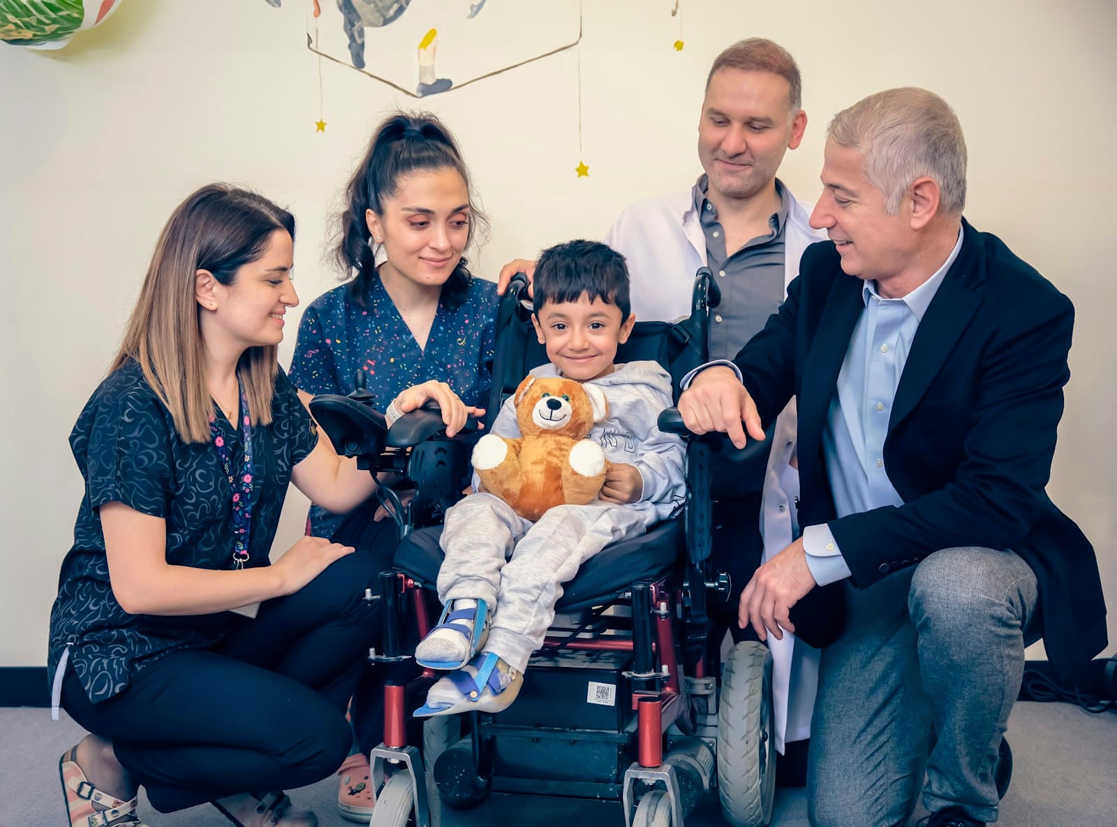 A young boy holding a stuffed bear smiles in his wheelchair, surrounded by four members of his care team.
