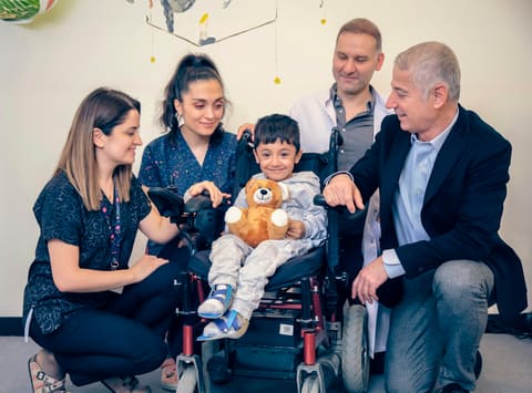 A young boy holding a stuffed bear smiles in his wheelchair, surrounded by four members of his care team.
