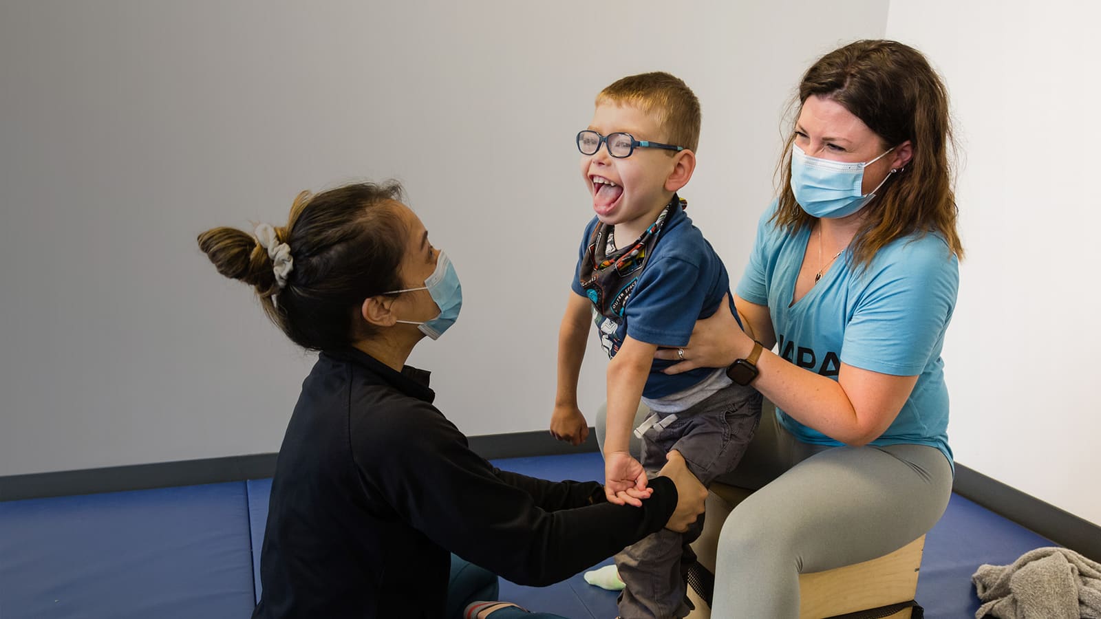 Two therapists support a laughing young child standing on a therapy mat.