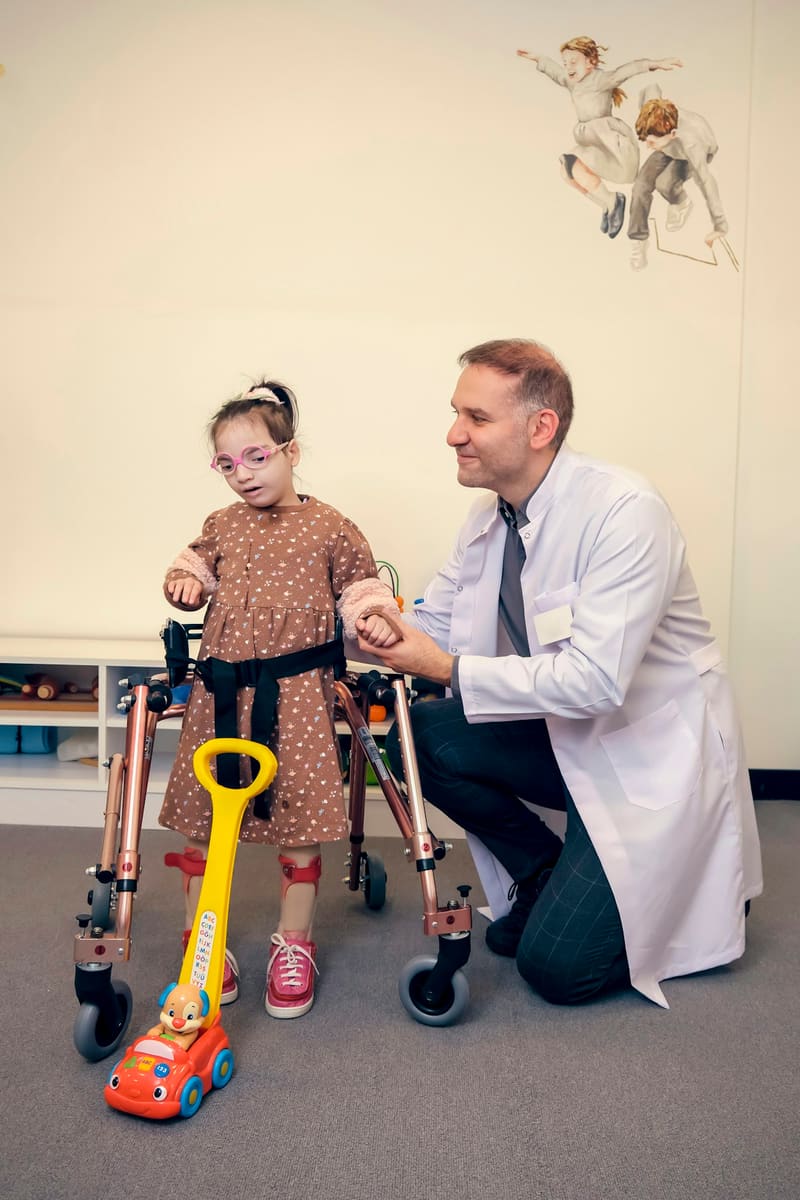 A young girl takes steps in a gait trainer while a clinician kneels beside her, holding her hand.