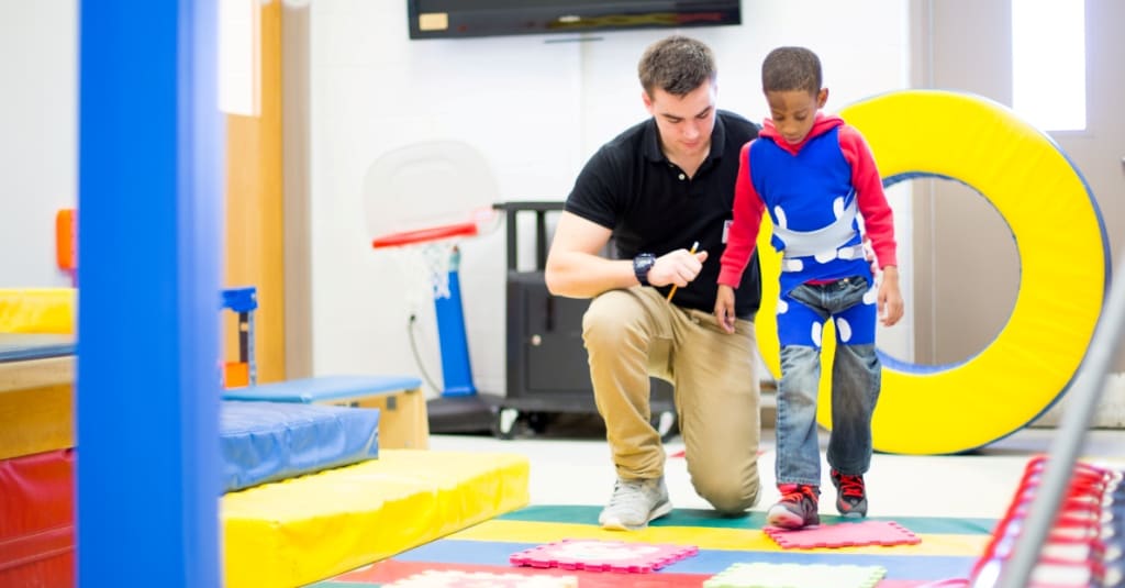 A physical therapist crouches beside a young child taking steps across a clinic gym floor.