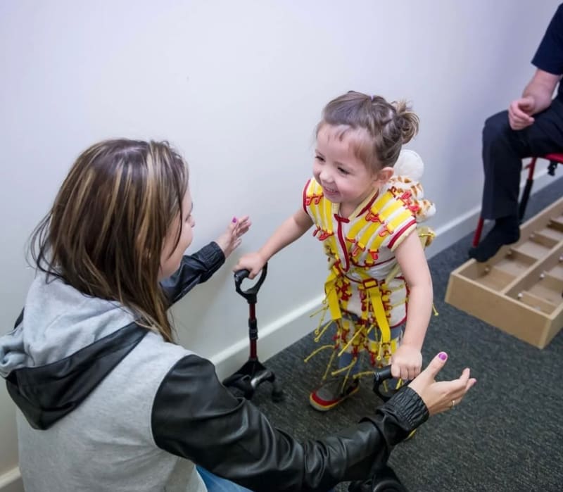 A young child in a support harness walks between handrails while an adult encourages them.