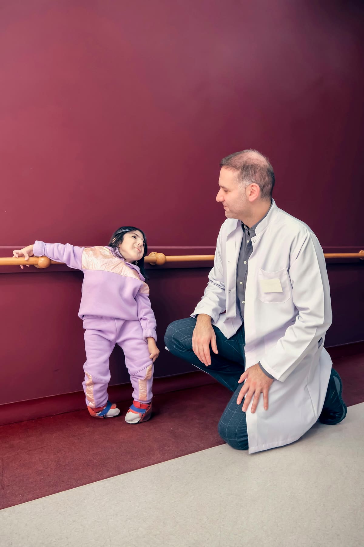A young girl in a supportive garment holds onto parallel bars during a standing exercise, with a clinician kneeling beside her.